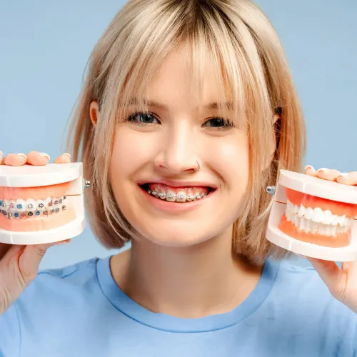 Smiling person wearing metal braces while holding two dental models, one with braces and one showing straight teeth to illustrate orthodontic treatment results.