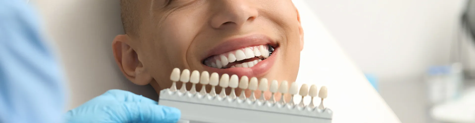 Gloved hand holding tooth shade-matching tool next to man's smile.