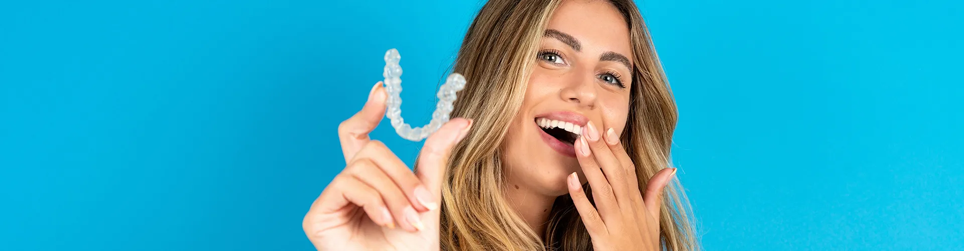 Smiling woman holding clear aligner and covering mouth.