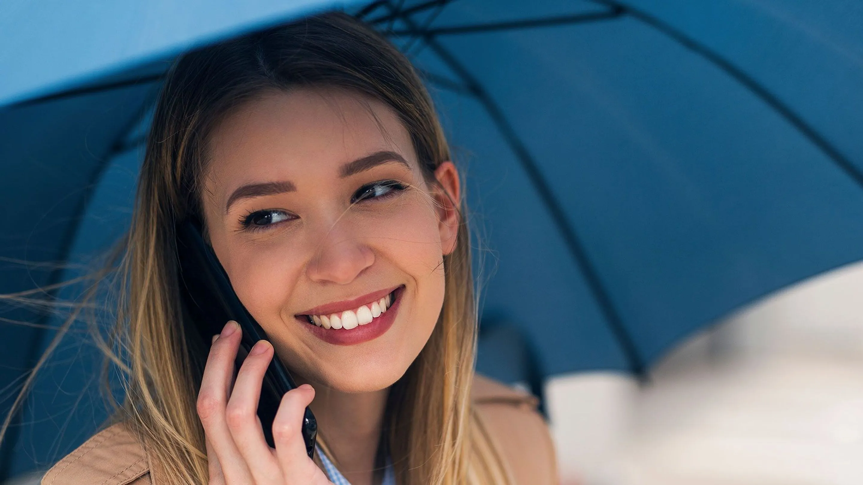 An image of a lady smiling while holding an umbrella while on the phone