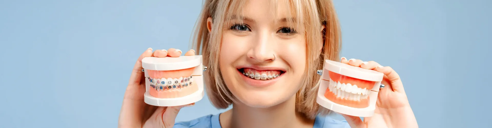 Smiling person wearing metal braces while holding two dental models, one with braces and one showing straight teeth to illustrate orthodontic treatment results.